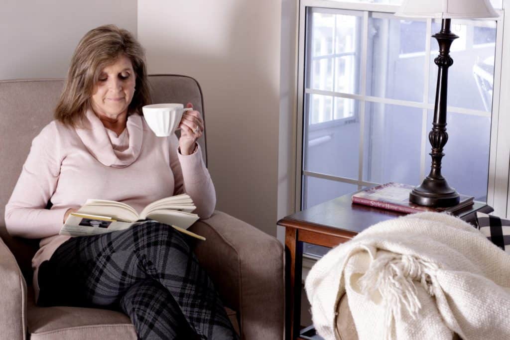 A woman holding a cup reading a book in a chair.