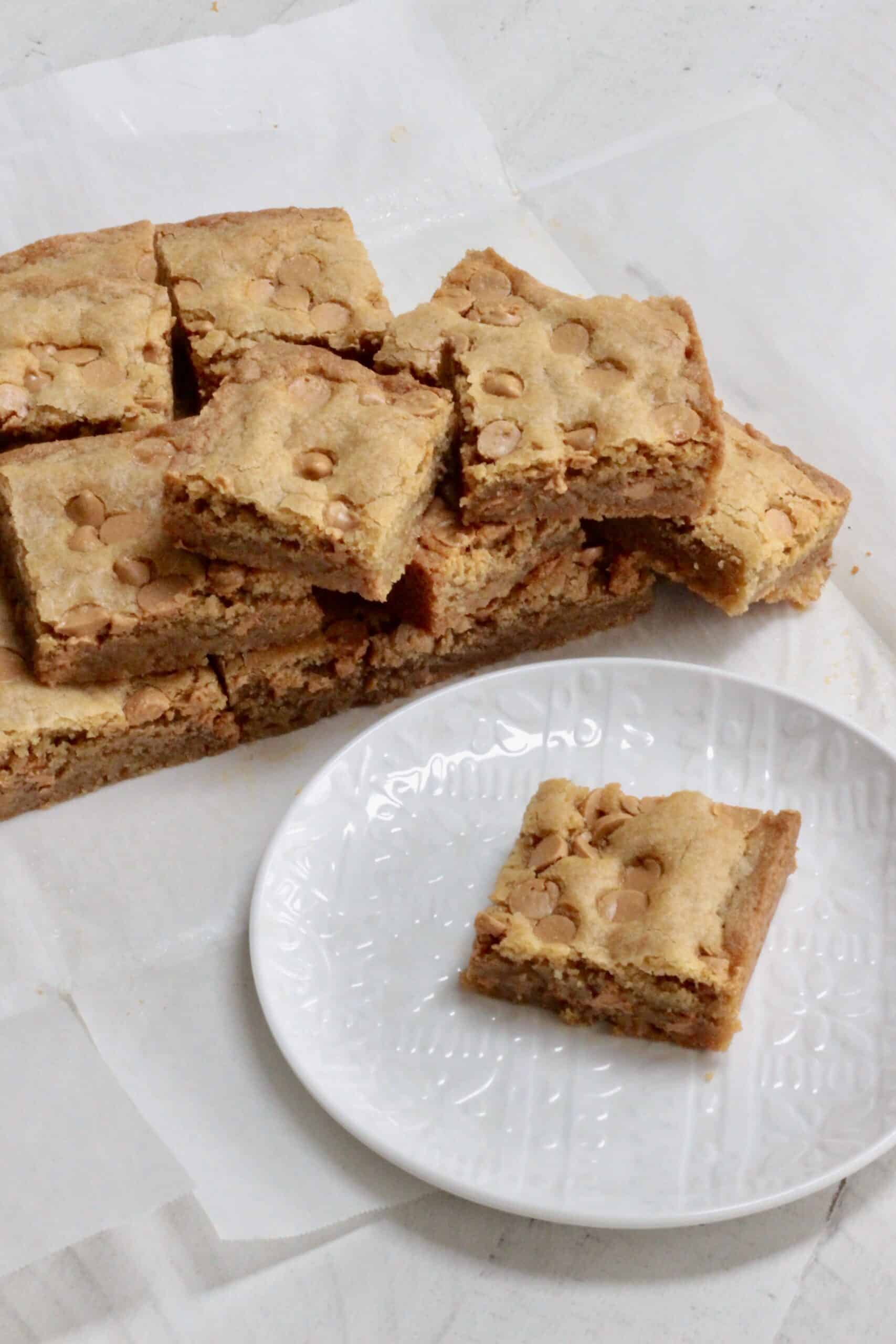 A stack of blondies in the background with one blondie on a plate.