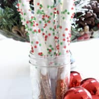 Almond bark dipped pretzels in a glass with red ornaments beside it.