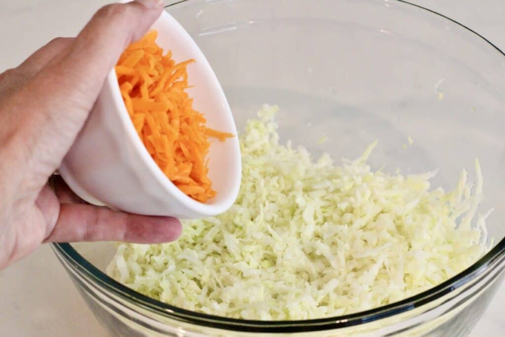 Adding grated carrots to a bowl of shredded cabbage.